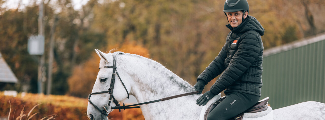 International showjumper Billy Twomey on the back of white horse while wearing GAIN jacket smiling at camera.